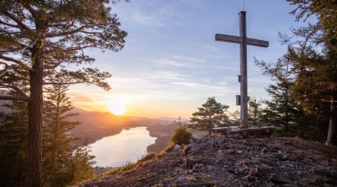 Holzkreuz auf einem Berggipfel mit Blick auf den Fuschlsee und Berge bei Sonnenuntergang, umgeben von Bäumen.
